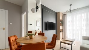 Uma sala de estar moderna com uma mesa de jantar redonda de madeira, cadeiras laranja, uma televisão de tela plana na parede e uma janela grande com cortinas leves.