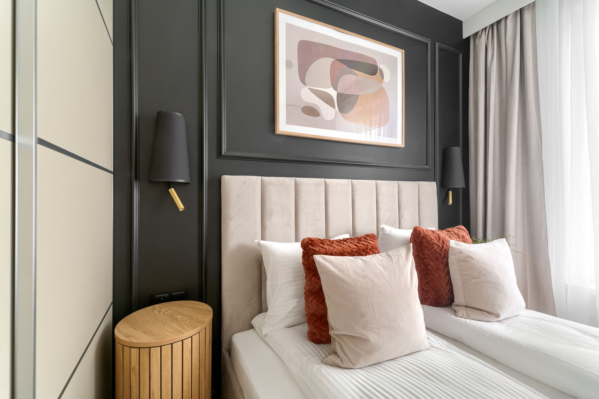 A modern hotel room with a large bed, a round wooden side table, and a framed abstract painting on the wall.
