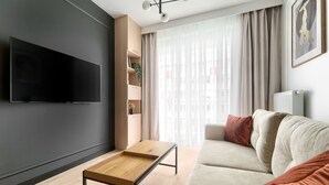 Uma sala de estar moderna com uma televisão de tela plana, uma mesa de centro de madeira, um sofá bege e uma janela com cortinas leves.
