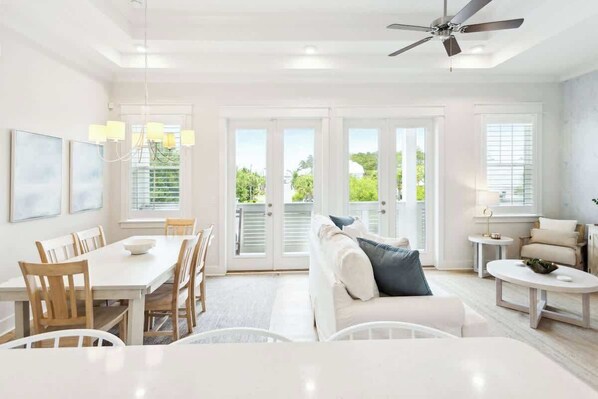 A bright, open living area with a dining table, white sofas, and a ceiling fan.