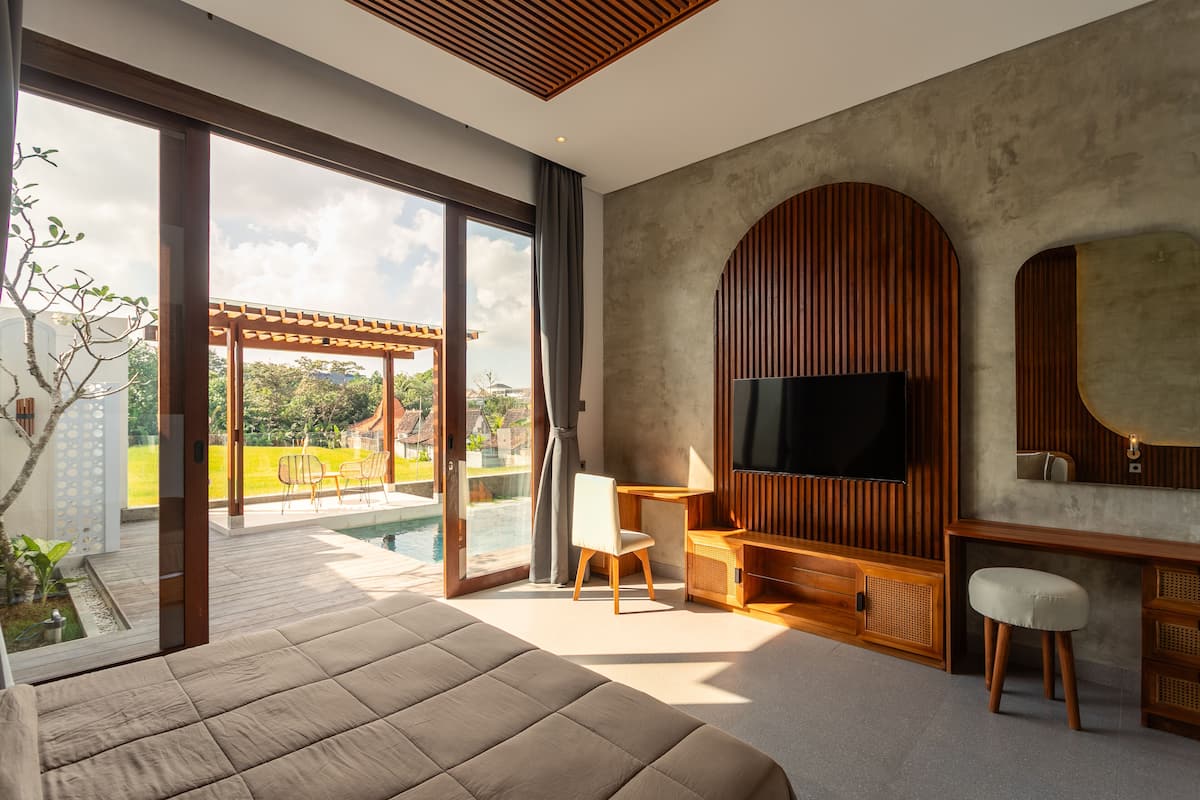 A modern bedroom with a large bed, a TV, a desk, and a view of a pool and garden.
