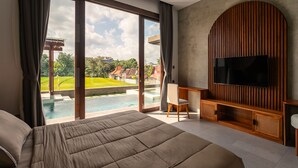 A bedroom with a large bed, a TV mounted on a wooden panel, a desk, and a view of a swimming pool and greenery outside. - Bellini Villa (Canggu)