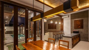 A modern dining area with a wooden table and chairs, a kitchen with bar stools, and a view of a swimming pool through glass doors. - Bellini Villa (Canggu)