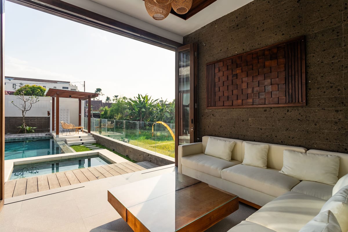A modern living room with a glass door leading to a pool area, a white sofa, and a wooden coffee table.