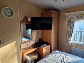 A compact room with a bed, a desk with a computer, a television mounted on the wall, a clock, and a window with curtains.