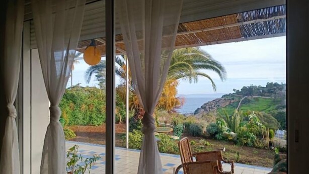 A room with a view of a coastal landscape, a wicker chair, and a potted plant. - Charmante Villa vue mer (Tanger)