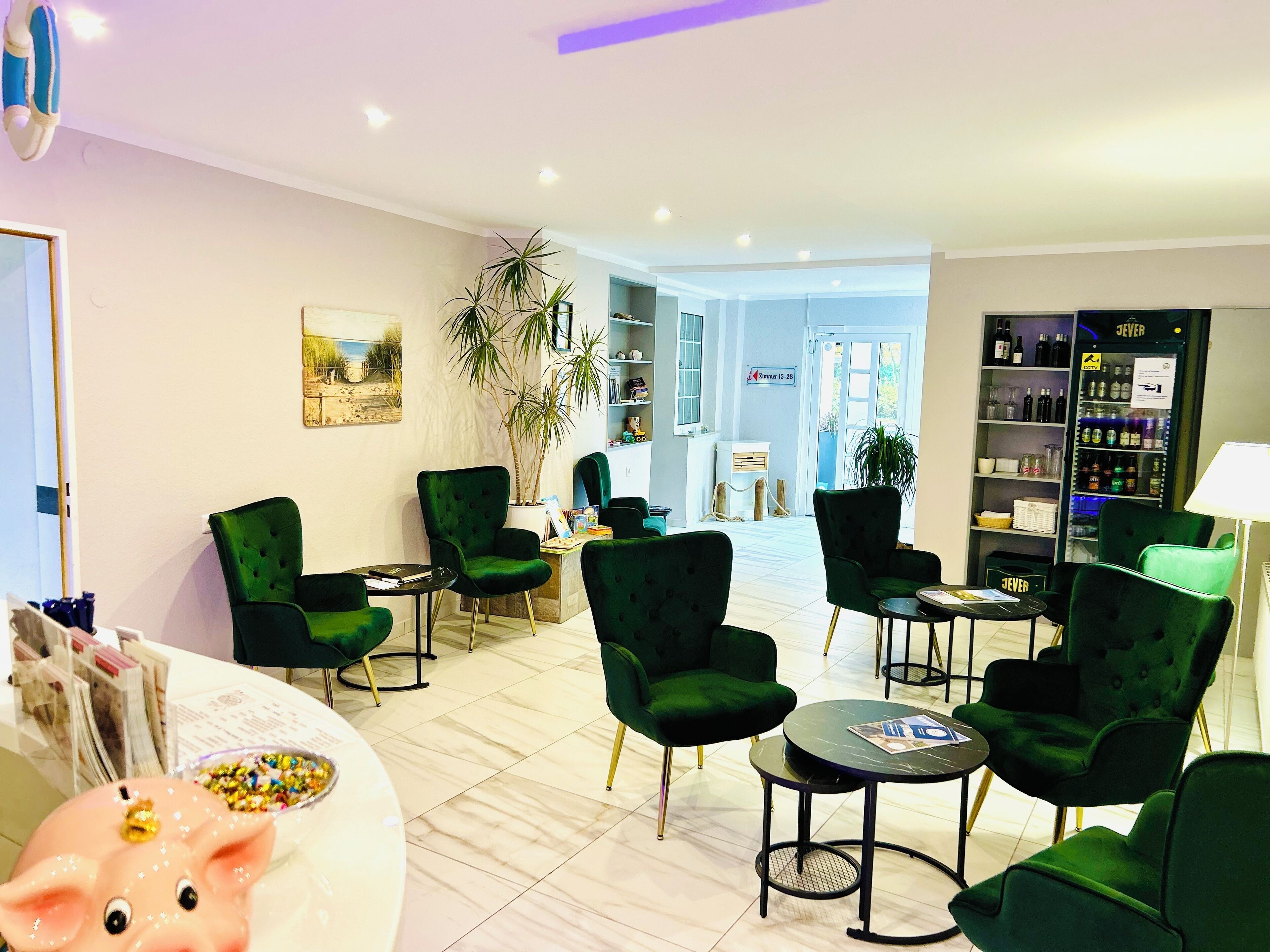 A modern lounge area with green upholstered chairs, a marble-tiled floor, a reception desk, and a wine cooler.