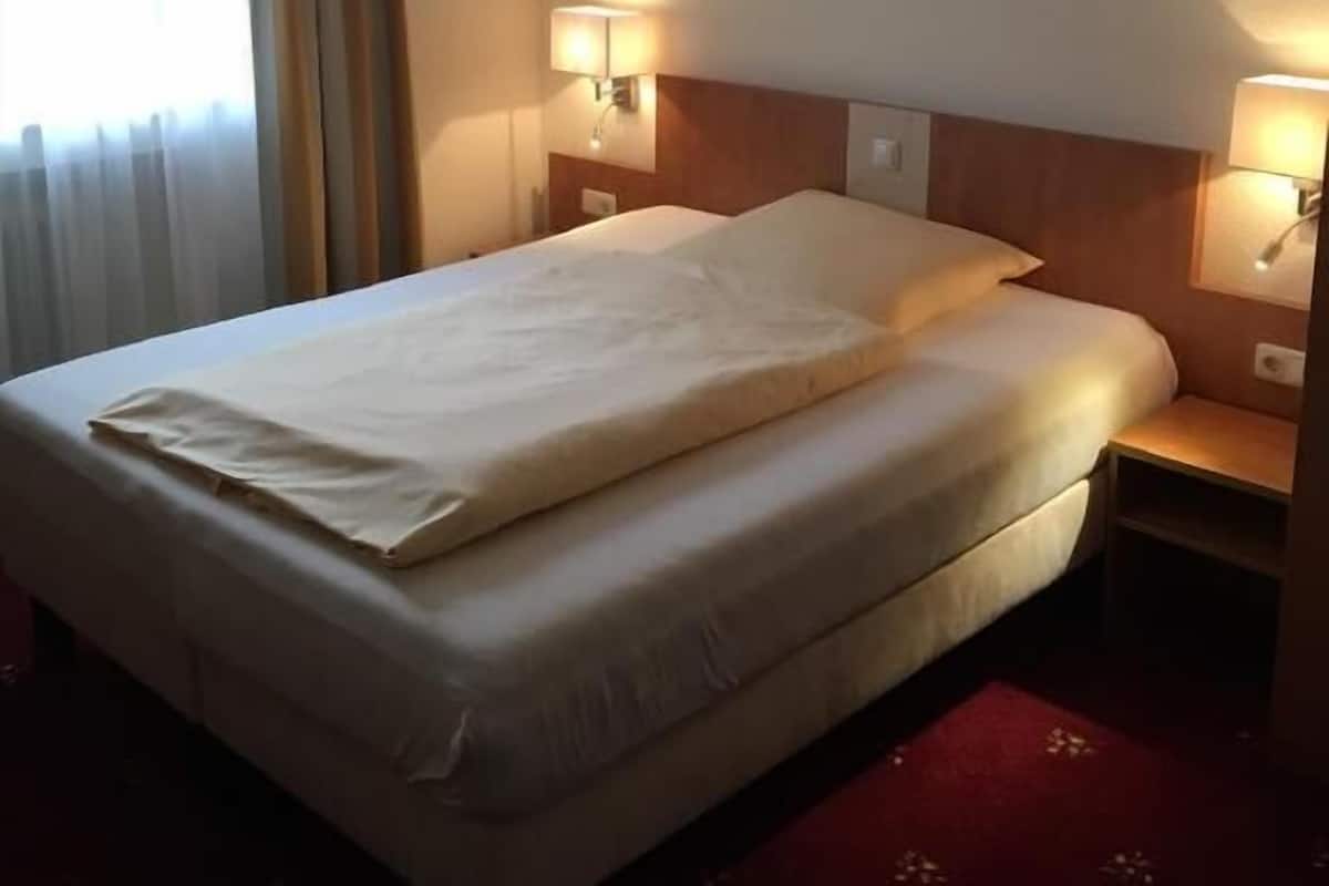 A hotel room with a bed, two bedside lamps, a window with curtains, and a red carpet.