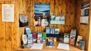 A wooden reception desk with informational posters, brochures, and a menu. - Tauwerk Hotel (Wilhelmshaven)