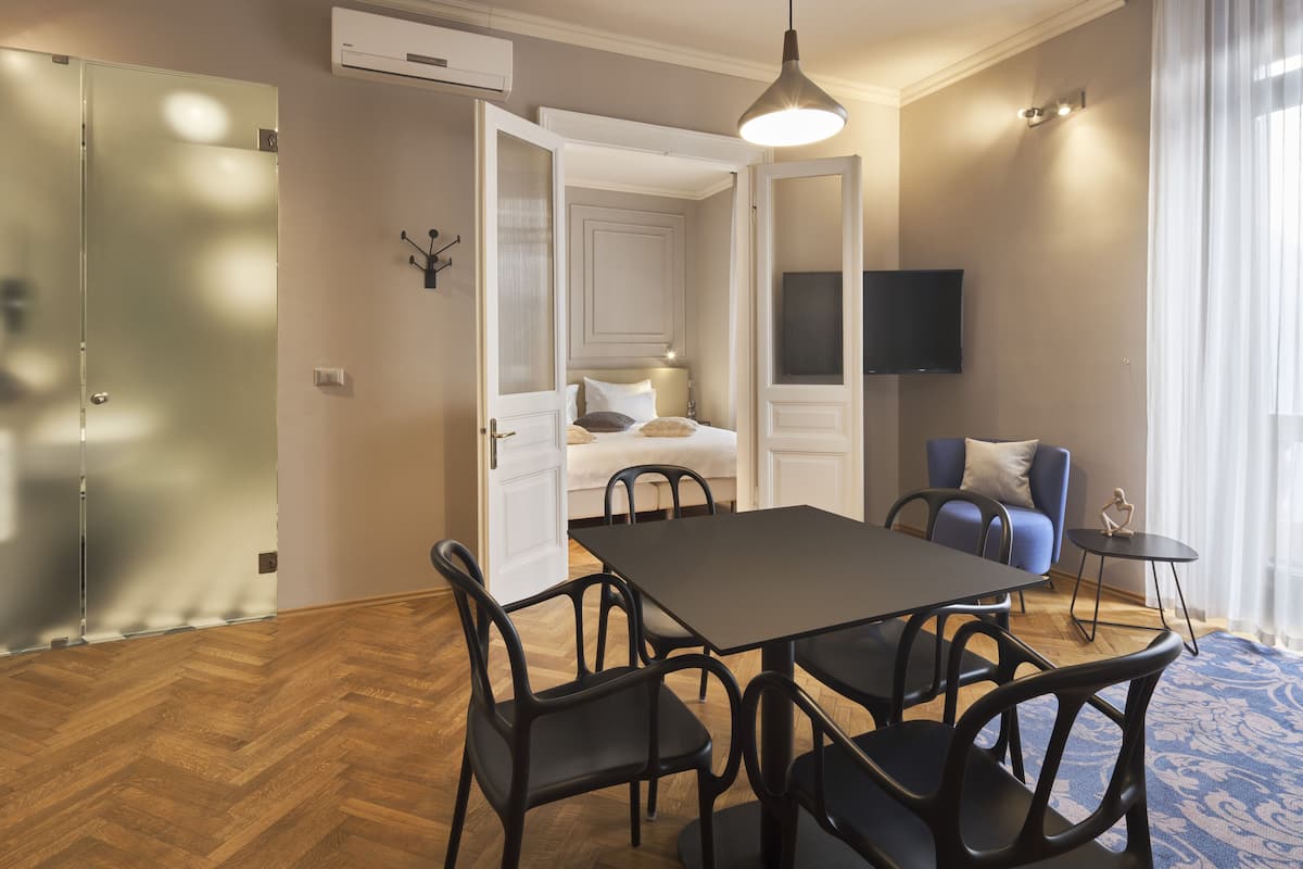 A modern hotel room with a dining area, a bed, a TV, and a view of the city.