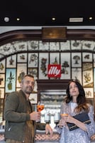 Due persone in piedi in un’area bar con una parete decorata da opere d’arte incorniciate e un cartello ‘illy’.