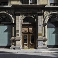 Facciata di un edificio con un'ampia porta in legno decorata, due finestre ad arco su ciascun lato e una targa con il numero civico.