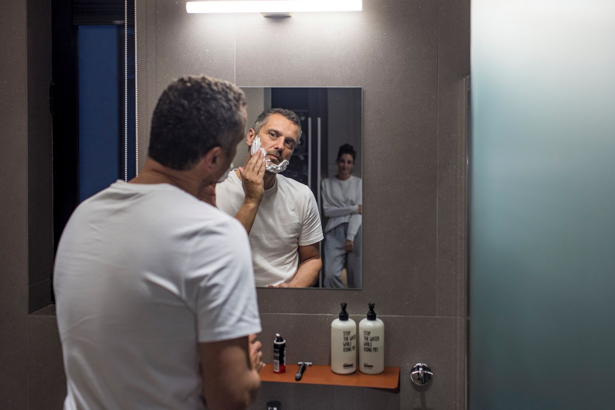 A man shaving in a bathroom with a mirror, a sink, and two bottles of soap on a shelf.