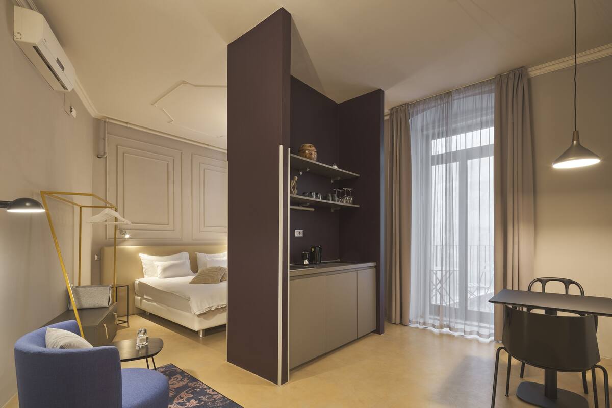 A compact hotel room with a kitchenette, a dining area, a sofa, and a large window with curtains.