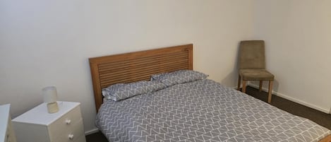 A single bed with a wooden headboard, a bedside table with a lamp, and a chair in a simple room with white walls.