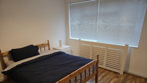 Une chambre avec un lit en bois, des tables de chevet blanches, une fenêtre avec des stores et un radiateur.