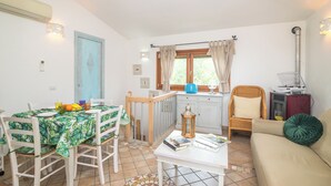 A dining area with a table set for four, a kitchen with a window, and a living area with a sofa and a lamp. - Villa del Golfo (Olbia)