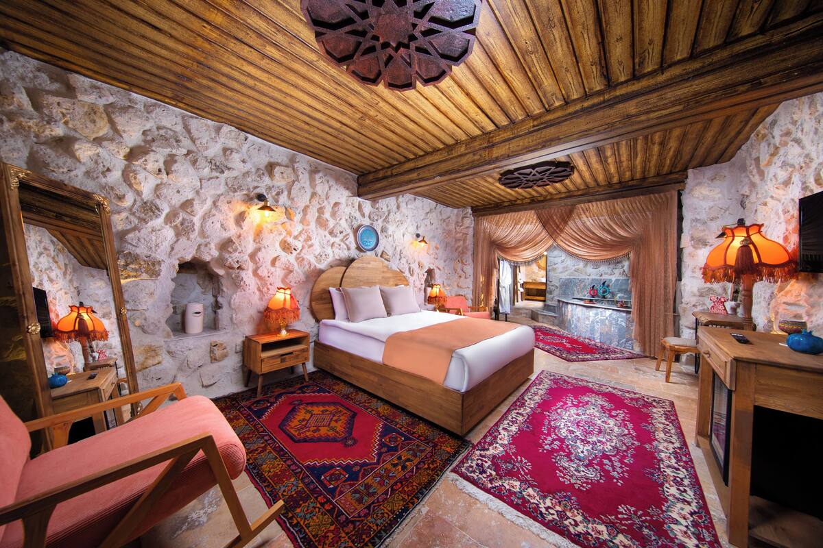 Un dormitorio con pared de piedra, techo de madera, una cama grande, dos mesitas de noche con lámparas, una silla y vistas a través de una ventana.