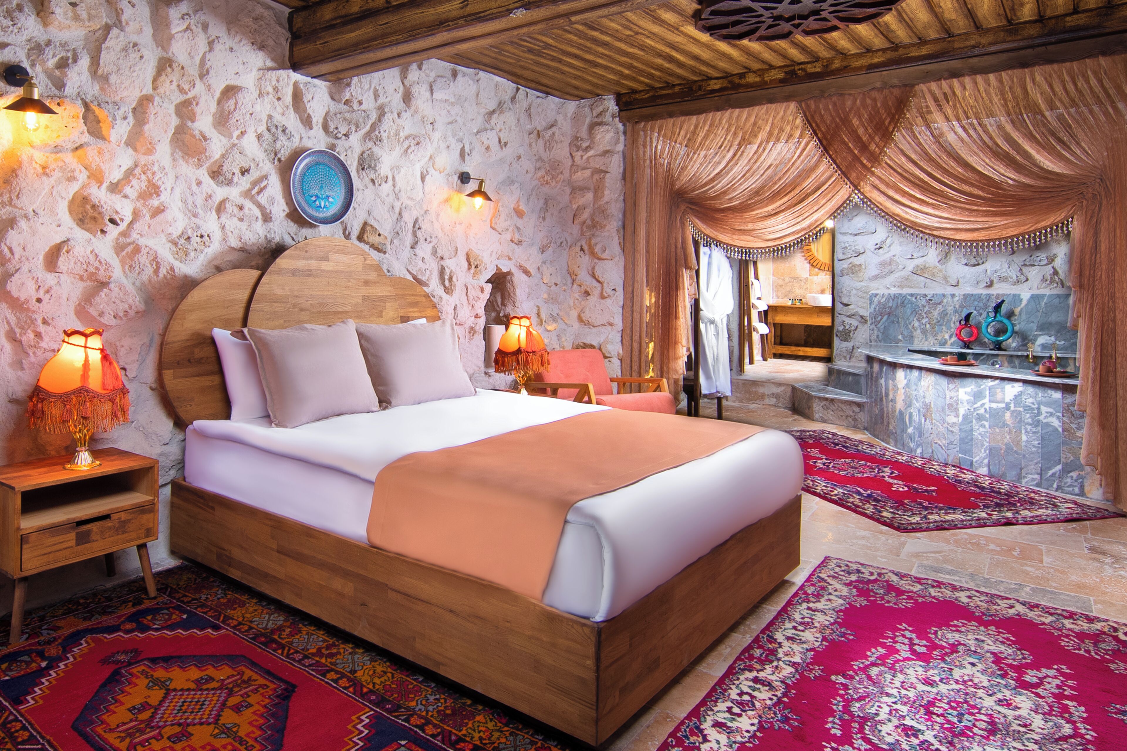 A bedroom with a stone wall, a wooden bed, two bedside lamps, a nightstand, and a bathroom visible through a window.