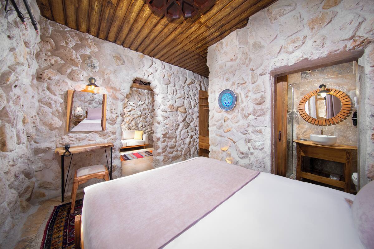 Una habitación de paredes de piedra con una cama, un escritorio de madera, un espejo y un baño con un espejo redondo y lavabo.