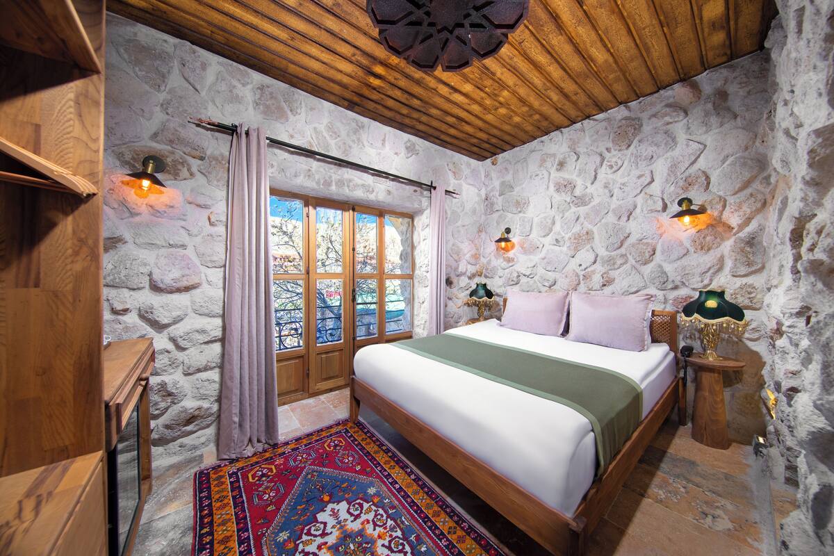 Un dormitorio con pared de piedra, techo de madera, una cama grande, dos lámparas de noche, un ventanal con cortinas y una alfombra colorida.