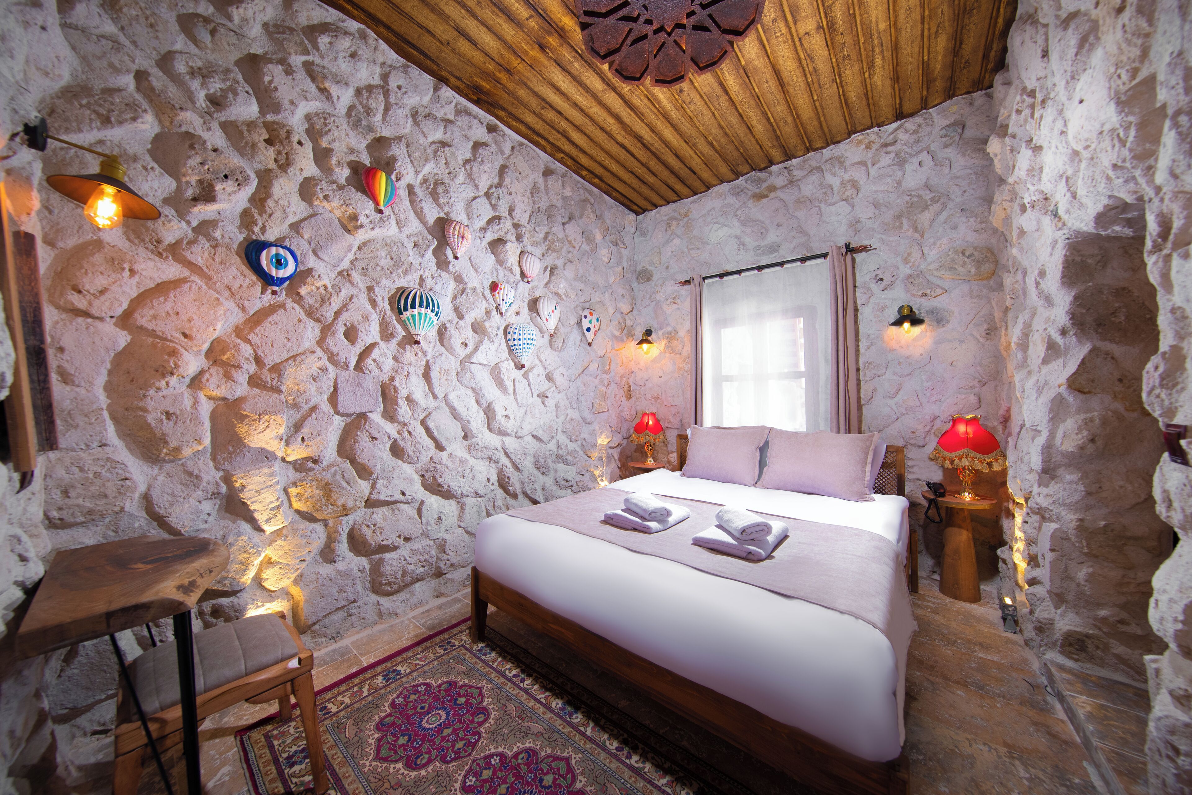 Habitación con paredes de piedra, techo de madera, una cama con toallas, una mesita y dos lámparas fijadas a la pared.