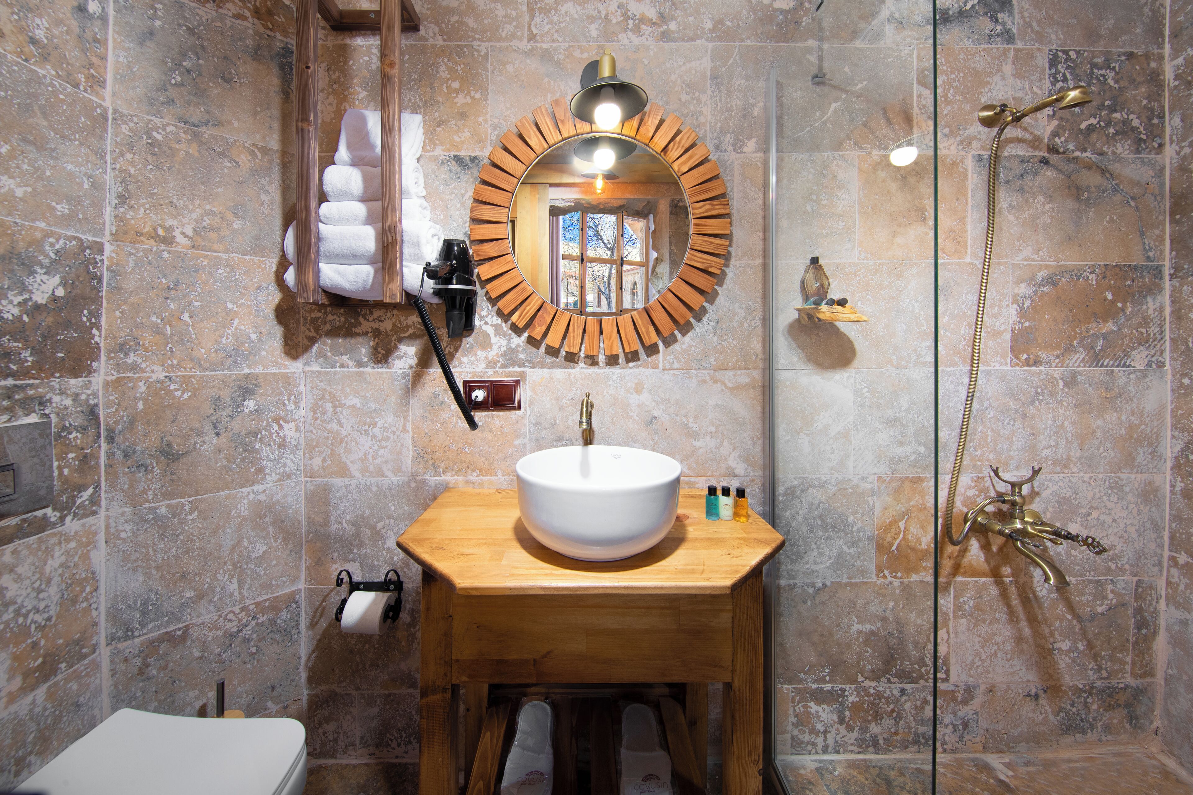 Un baño con tocador de madera, un espejo redondo, un lavabo blanco y una ducha con cabezal dorado.