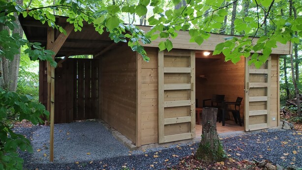 Une cabane en bois, avec un sol recouvert de gravier, une porte, et une petite table et une chaise à l’intérieur.