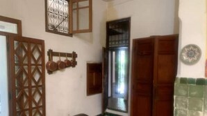 A room with a wooden door, a window with a metal grill, a wall-mounted shelf with pots, and a tiled floor. - DAR Benjelloun Studio Plat (Fès)