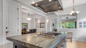 Una cocina moderna con una isla grande, electrodomésticos de acero inoxidable, iluminación colgante y vistas al exterior a través de un ventanal.