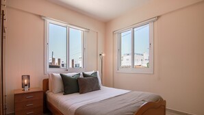 A bedroom with a bed, nightstand, lamp, and large windows. - Phaedrus Living: Universal City Flat Alina (Paphos)
