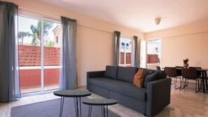 A living room with a sofa, two small tables, a dining area with chairs, and a sliding glass door leading to an outdoor area. - Phaedrus Living: Universal City Flat Alina (Paphos)