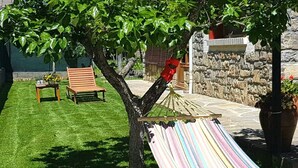 Un hamac est attaché à un arbre dans un jardin bien entretenu, avec un mur de pierres et un banc.