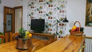 Un coin repas comprenant une table en bois, des chaises, un téléviseur, un panier de fruits et un mur décoré d’un motif de fruits.