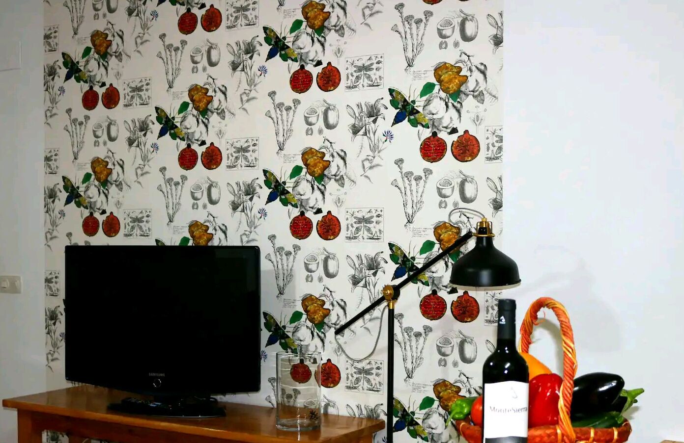 Een kamer met een televisie op een houten standaard, een zwarte bureaulamp, een fles wijn en een mand met groenten.