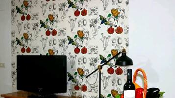Een kamer met een televisie op een houten standaard, een zwarte bureaulamp, een fles wijn en een mand met groenten.