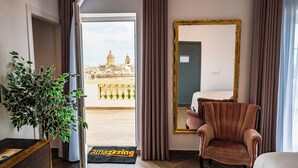 A hotel room with a large mirror, a plush armchair, a potted plant, and a balcony with a city view. - Yolanda Boutique Hotel  (Birgu)