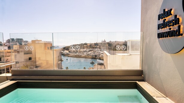 Piscina en la azotea con vista a la ciudad, un letrero que dice 'relájate, refréscate, desconecta' y símbolos de prohibido fumar en el vidrio.
