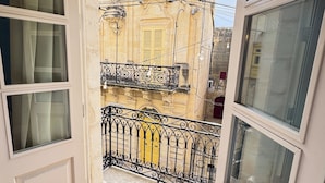 A view through an open door to a balcony with a wrought iron railing, a yellow door, and a building facade with classical architecture. - Mosta Stays  (Mosta)