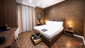 A modern hotel room with a large bed, wooden flooring, a desk with a chair, a small table with a lamp, and a wall-mounted TV.
