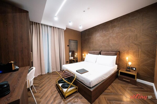 A modern hotel room with a large bed, wooden flooring, a desk with a chair, a small table with a lamp, and a wall-mounted TV.