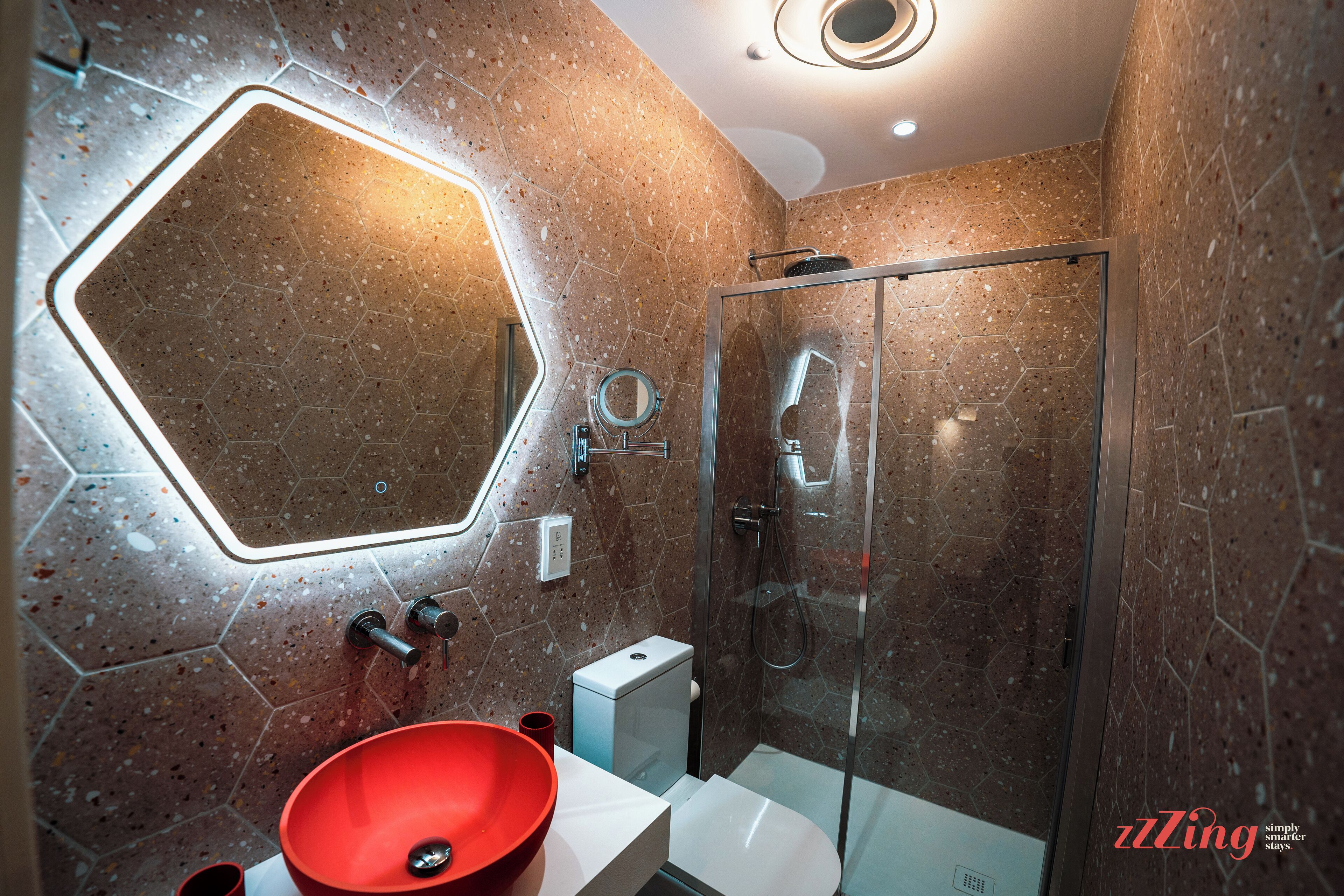 A modern bathroom with a hexagonal mirror, a glass shower enclosure, and a red basin.