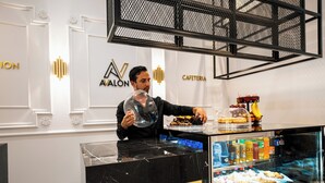 A person serving food at a counter with a display case of pastries and a beverage cooler.
