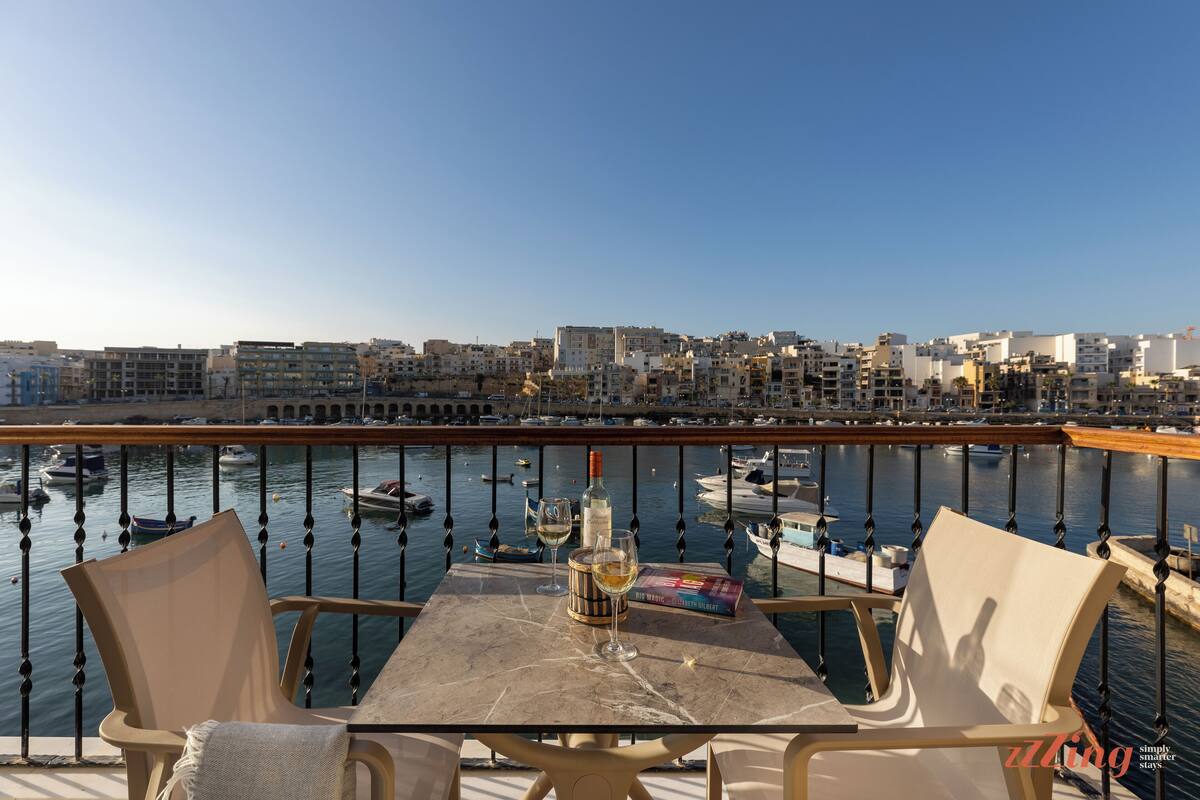 Un balcone con tavolo, due sedie, una bottiglia di vino e un bicchiere. Si vede un porto turistico con barche e uno skyline sullo sfondo.
