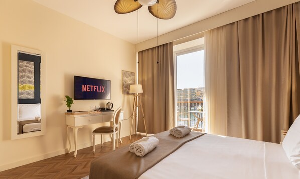 A hotel room with a bed, a desk, a chair, a TV displaying 'NETFLIX', and a balcony with curtains.