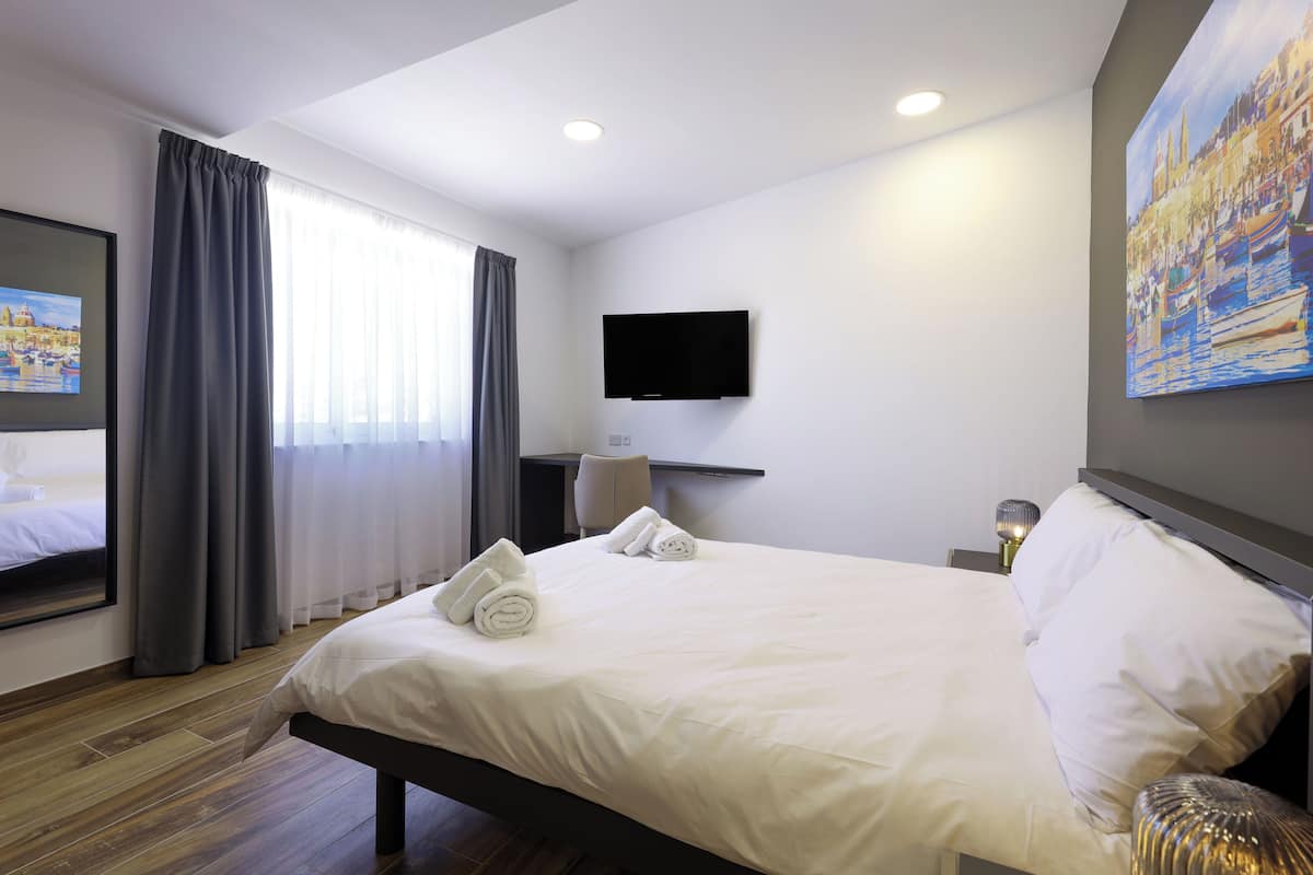 Ein Hotelzimmer mit einem großen Bett, einem an der Wand befestigten Flachbildfernseher, einem Stadtbildgemälde und einem Vorhangfenster.