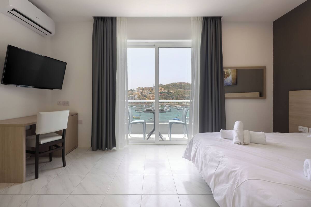 Ein Hotelzimmer mit Bett, Schreibtisch, Stuhl, Fernseher und einem Balkon mit Meerblick, sowie einem Bild an der Wand.