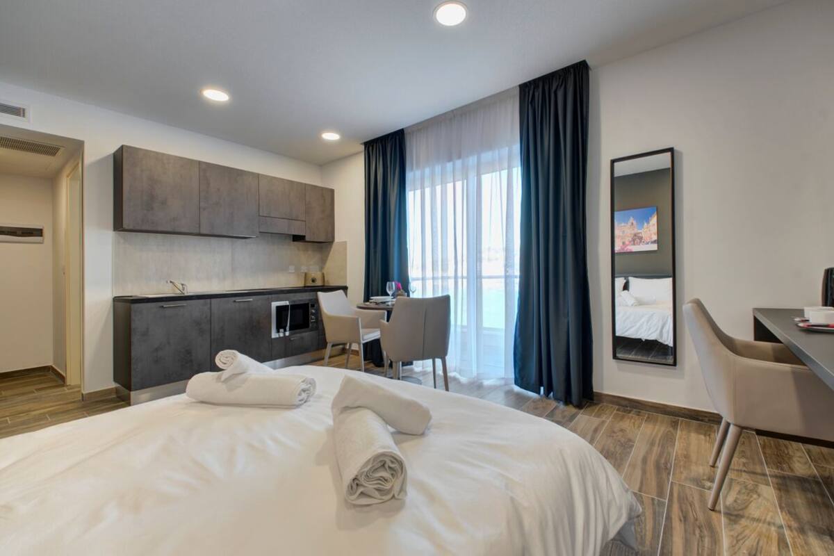 Ein Hotelzimmer mit Bett, Schreibtisch, Stuhl, einer Küchenzeile und einem großen Fenster mit Vorhängen.