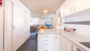 Ett modernt kök med vita skåp, en matplats med ett bord för två personer och ett vardagsrum med en soffa och en tv.