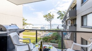 Un balcon avec un barbecue, des sièges d’extérieur, une table avec du vin et des raisins, et une vue sur la mer et les bâtiments.